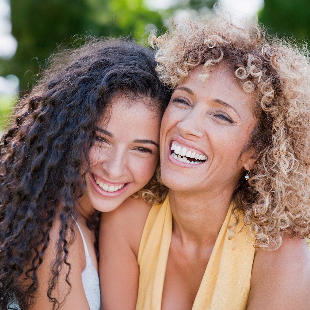 Close up of two female best friends laughing and embracing each other. They each have beautiful curly hair, one has long brunette 2C curls, while the other fashions shoulder length, blonde, 3B curls.